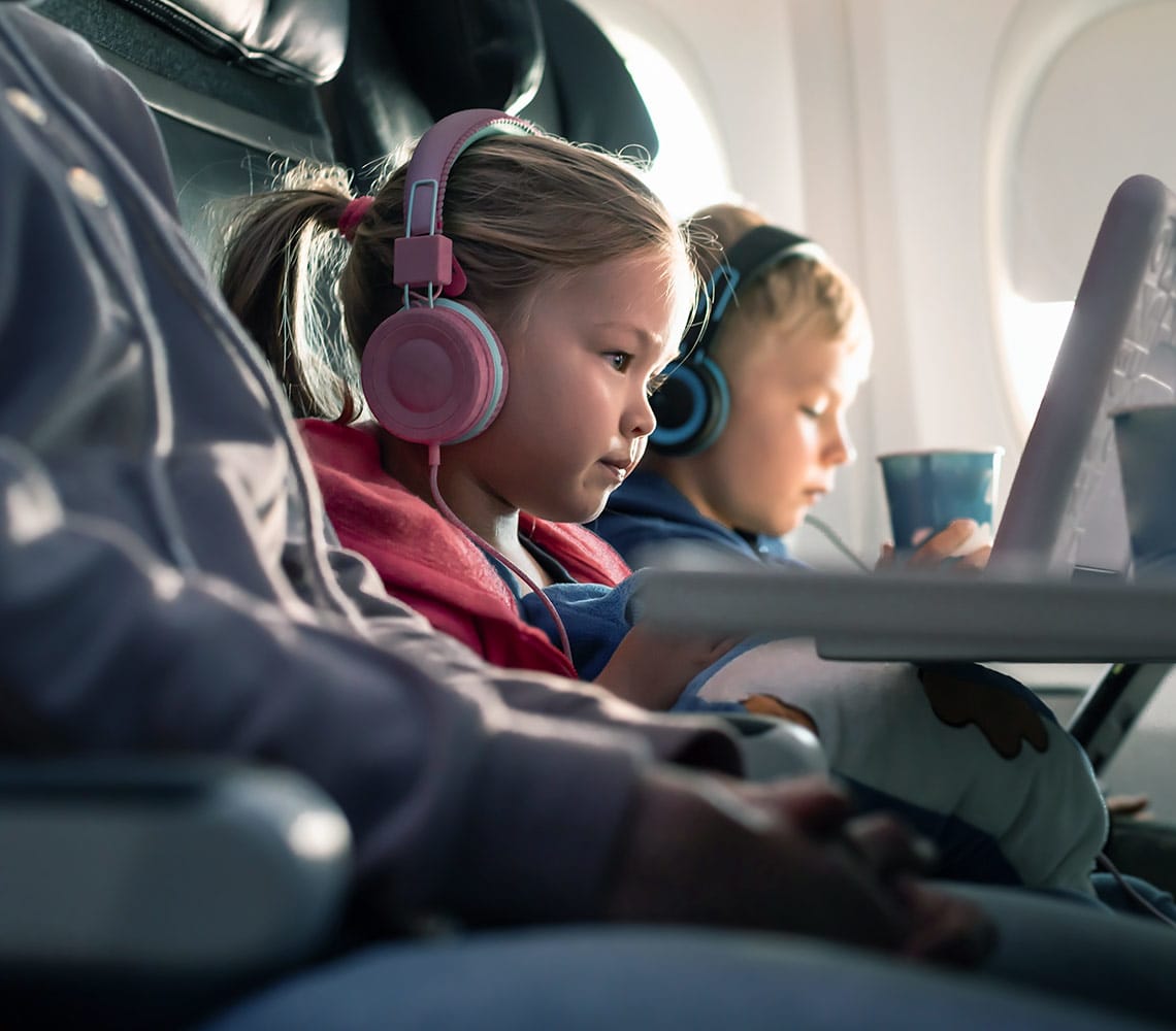 Two children, a girl and a boy, sitting on an airplane with headphones on, engaged in their in-flight entertainment.